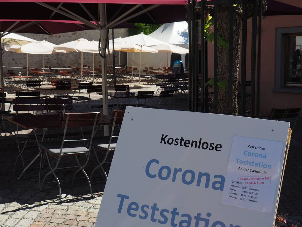 Nahaufnahme eines Informationsschilds zur Teststation an der Eselsmühle sowie Außengastronomiebestuhlung und der Teststation im Hintergrund