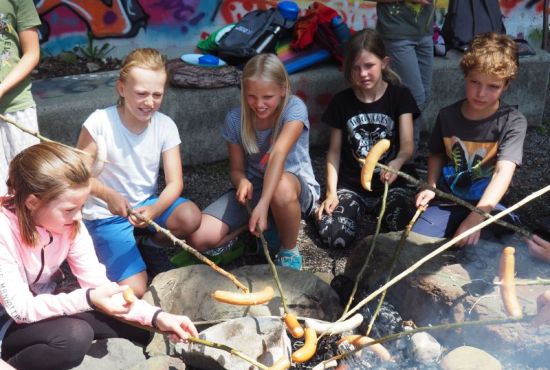 Mehrere Kinder sitzen um ein Lagerfeuer und halten Würstchen an Stöcken übers Feuer