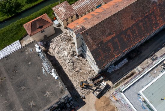 Blick von oben auf zwei alte Fabrikgebäude, dazwischen steht ein Bagger und schaufelt Schutt in einen bereit stehenden Lastwagen