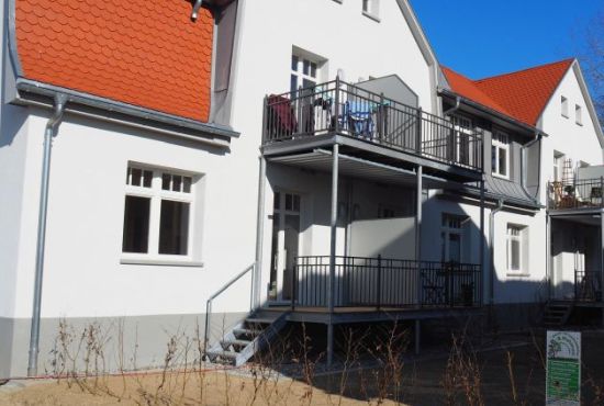 Blick auf ein saniertes Wohngebäude mit roten Dachziegeln und vorgestzten Stahlbalkonen im oberen und unteren Stock