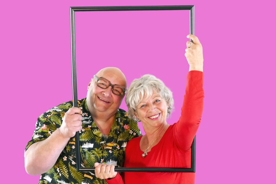 Ein Mann und eine Frau schauen durch einen Bilderrahmen und lächeln fröhlich in die Kamera.