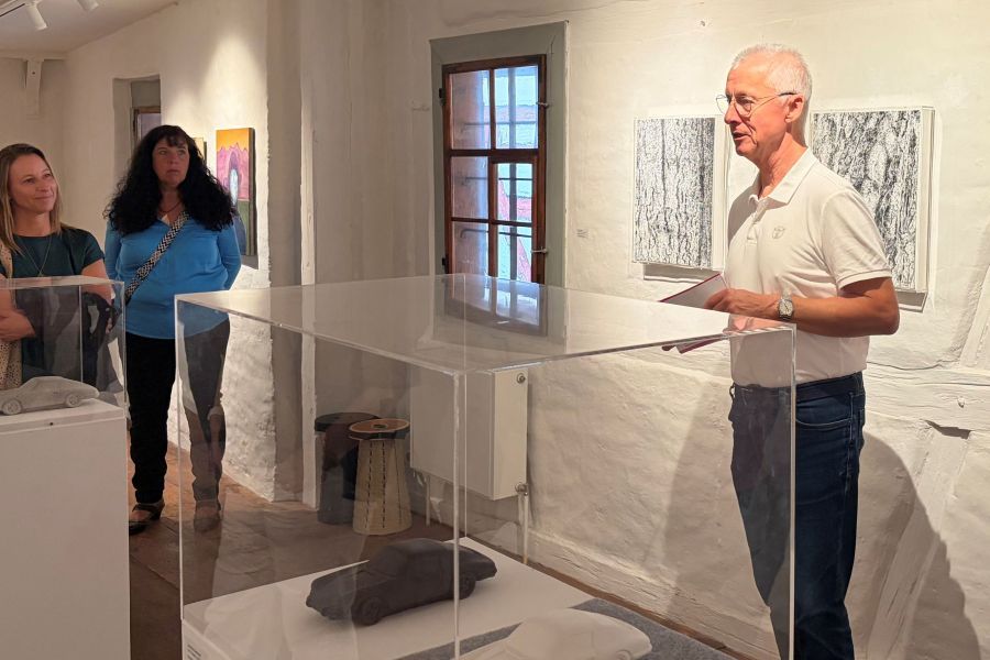 Eine Person präsentiert in einem Ausstellungsraum Kunstwerke. Zwei weitere Personen hören aufmerksam zu. Der Raum hat helle Wände und ist mit Kunstwerken dekoriert. Eine transparente Vitrine steht im Vordergrund, in der sich ein graues Objekt befindet.