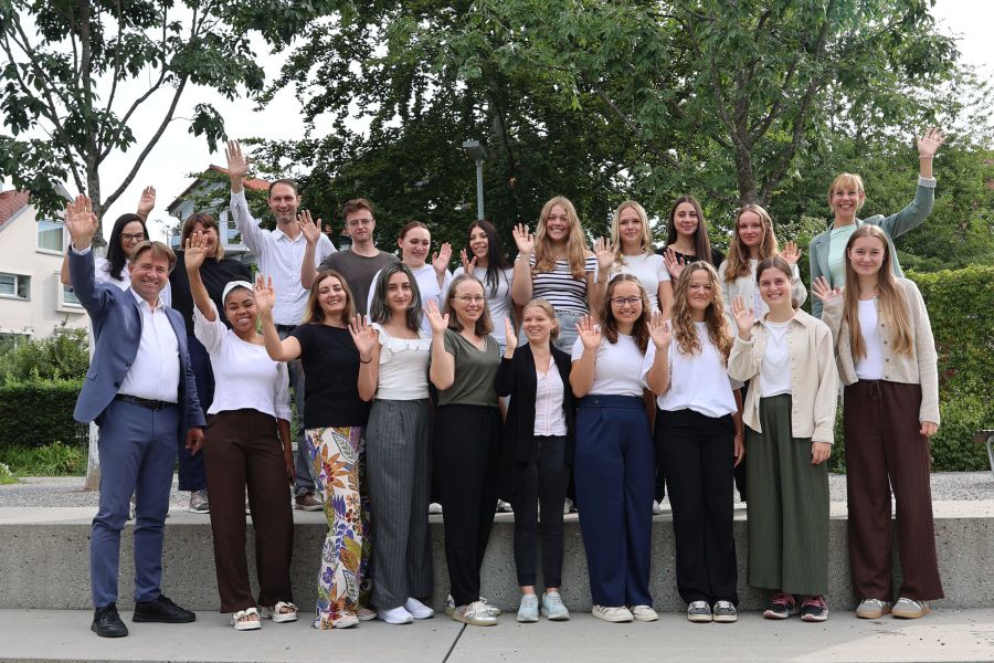 Eine Gruppe von etwa 20 Personen steht im Freien und winkt in die Kamera. Die Personen sind unterschiedlich gekleidet und lächeln. Im Hintergrund sind Bäume und Gebäude zu sehen, die eine freundliche Atmosphäre schaffen.