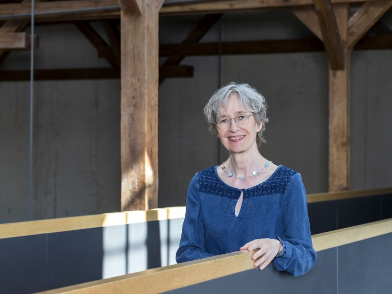 Eine Frau mit kurzen, grauen Haaren steht an einem Holzbalkon. Sie trägt ein blaues Oberteil mit einem dekorativen Ausschnitt und eine Kette. Im Hintergrund sind Holzstützen und eine schlichte Wand zu sehen. Die Frau lächelt freundlich in die Kamera.