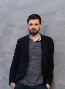 Ein Mann mit Bart steht an einer grauen Wand. Er trägt ein schwarzes Jacket und ein gestreiftes Hemd. Der Mann schaut direkt in die Kamera und hat eine entspannte Haltung.