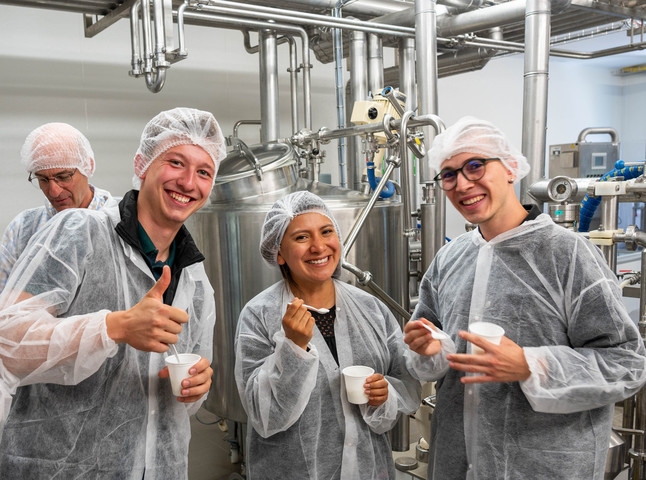 Eine Gruppe von vier Personen in Schutzkleidung steht in einer industriellen Umgebung. Drei Personen lächeln und geben Daumen hoch, während eine Person im Hintergrund zuschaut. Alle halten kleine Becher in der Hand. Im Hintergrund sind Maschinen und Rohre zu sehen.