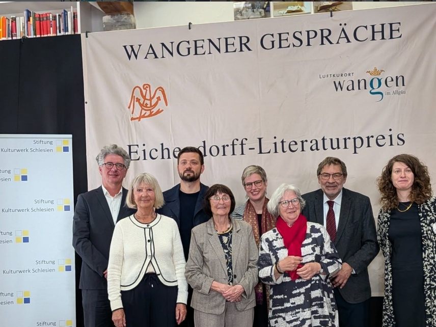 Eine Gruppe von sieben Personen steht vor einem Banner mit der Aufschrift "Wangener Gespräche" und "Eichendorff-Literaturpreis". Die Personen lächeln und sind in formeller Kleidung gekleidet. Im Hintergrund sind Regale mit Büchern zu sehen.