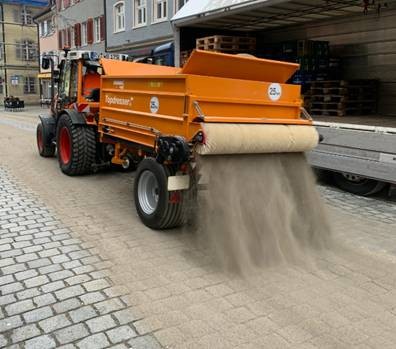 Fahrzeug des Bauhofs verteilt Sand auf dem Pflaster 