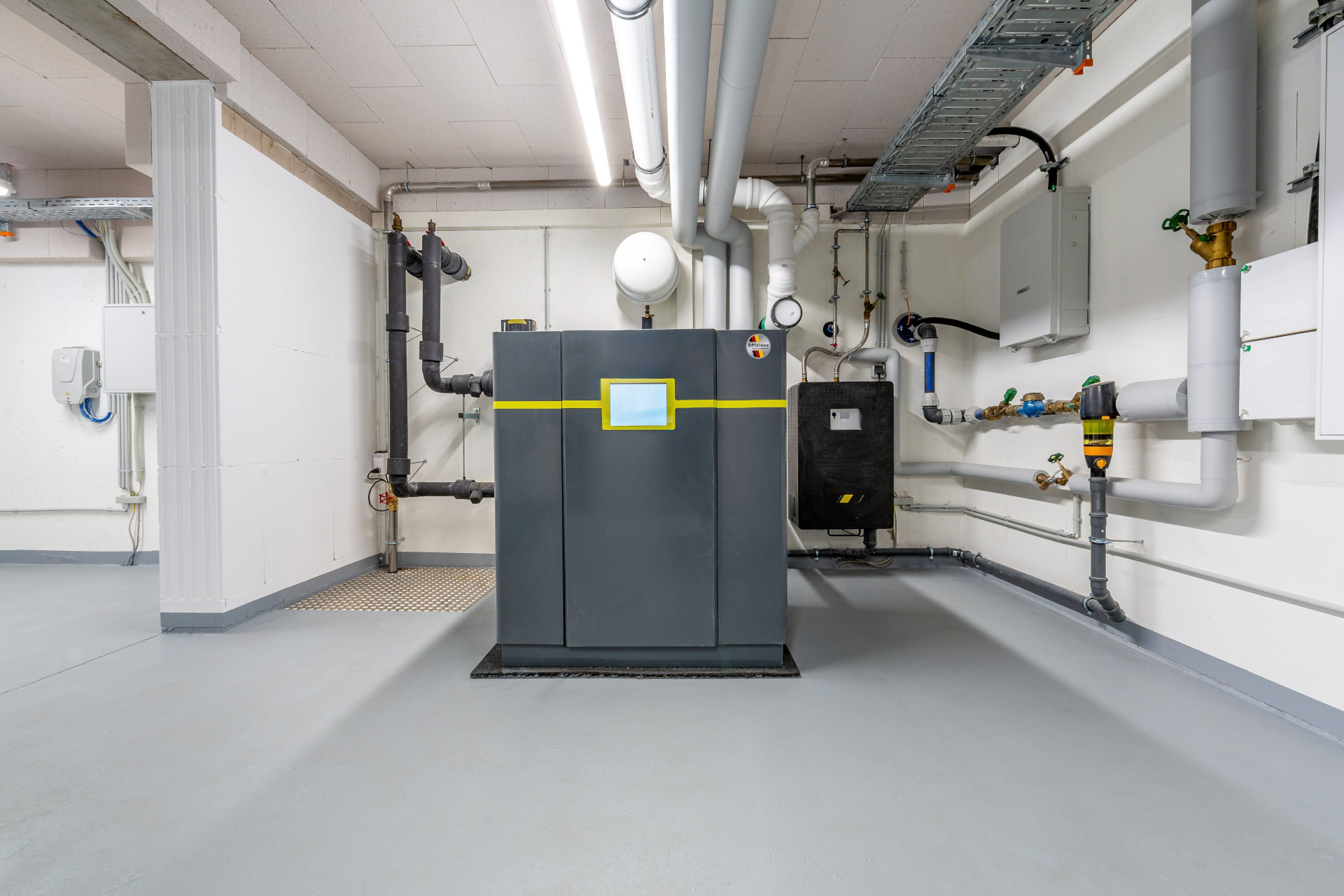 Ein moderner Heizungsraum mit einer großen Heizungsanlage in der Mitte. Um die Anlage verlaufen Rohre, und an den Wänden sind verschiedene technische Geräte und Anschlussstellen sichtbar. Der Boden ist grau und der Raum gut beleuchtet.