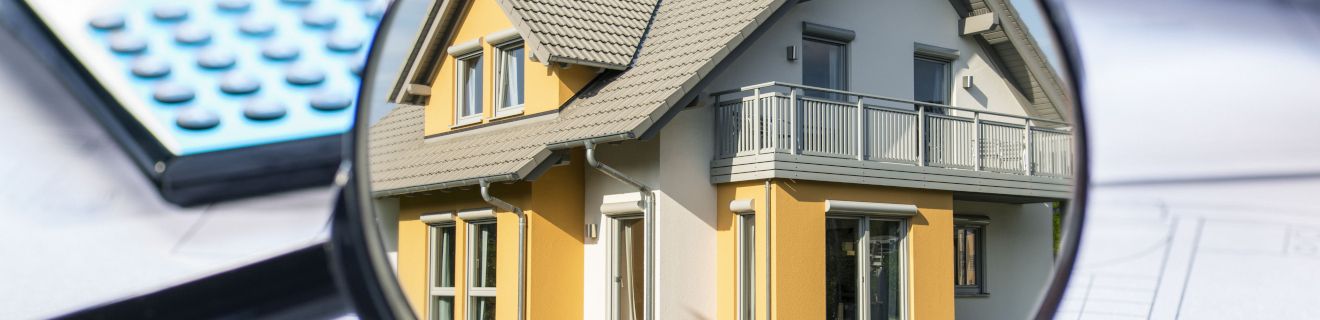 Ein modernes Haus mit gelber Fassade und Balkon, das durch eine Lupe betrachtet wird. Im Hintergrund sind Planungsunterlagen und ein blauer Taschenrechner sichtbar, die den Kontext von Gebäudekosten oder Immobilienbewertung andeuten.