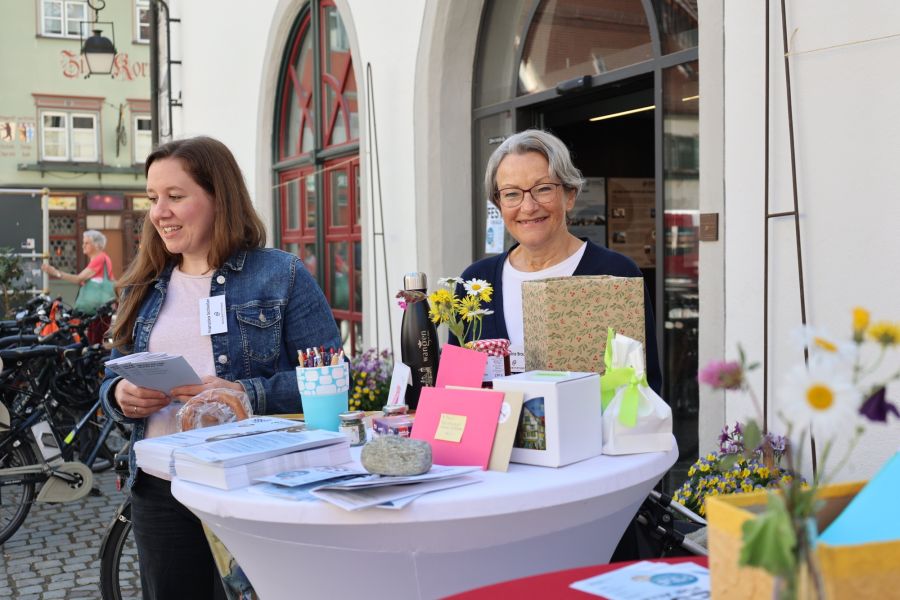 Zwei Frauen stehen an einem Tisch im Freien, umgeben von Geschenken und Blumen. Eine Frau hat ein Heft in der Hand und lächelt, während die andere freundlich in die Kamera schaut. Im Hintergrund sind Fahrräder und ein Gebäude sichtbar.