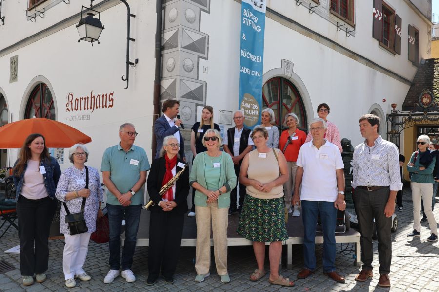 Eine Gruppe von Menschen steht auf einer Bühne vor einem Gebäude mit dem Schriftzug "Rornhaus". Sie lächeln und posieren für die Kamera, während ein Mann am Mikrofon spricht. Im Hintergrund ist ein Banner mit Informationen zu einer Veranstaltung.