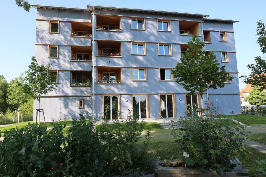 Ein modernes, mehrstöckiges Wohngebäude mit blauer Fassade und Holzelementen. Es verfügt über Balkone mit Blumenarrangements. Vor dem Gebäude gibt es einen gepflegten Garten mit verschiedenen Pflanzen und Bäumen, sowie einen Zugang mit Treppen.