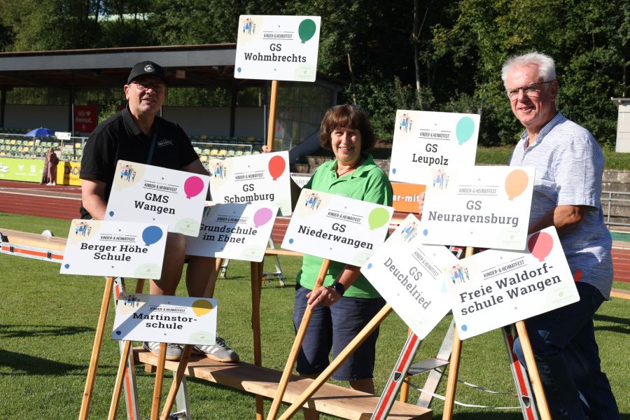 Drei Personen stehen auf einem Sportplatz zwischen Schildern mit Schulnamen. Die Schilder repräsentieren verschiedene Schulen, darunter GMS Wangen, GS Leupolz und Freie Waldorfschule Wangen. Die Szene vermittelt eine freundliche Atmosphäre im Rahmen eines Festes oder einer Veranstaltung.