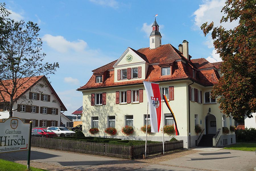 Das Rathaus in Leupolz mit roten Fensterläden sowie davor eine Deutschlandfahne und eine Fahne mit dem Wangener Wappen