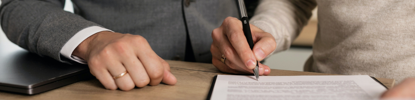 Zwei Menschen an einem Tisch, von denen man nur die Hände sieht, eine der beiden Personen unterschreibt ein Formular