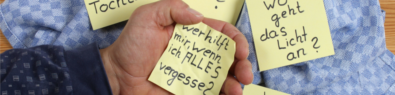 Eine Hand hält einen gelben Zettel mit der Frage: "Wer hilft mir, wenn ich ALLES vergesse?" Um die Hand herum liegen weitere gelbe Zettel mit verschiedenen Fragen. Der Hintergrund besteht aus einem blauen Tuch und einem Holztisch.