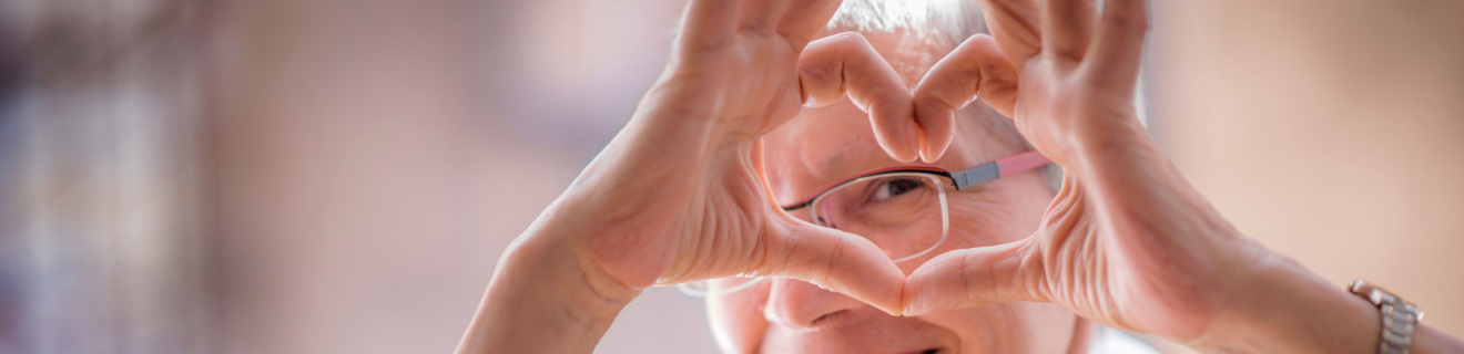 Eine ältere Person formt mit ihren Händen ein Herzsymbol, lächelt und trägt eine Brille. Der Hintergrund ist unscharf und zeigt warme Farben.