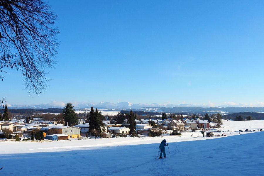 Ein Kind fährt auf der Piste des Skilifts Berger Höhe. Im Hintergrund ist ein Wohngebiet und am Horizont die Alpen zu sehen