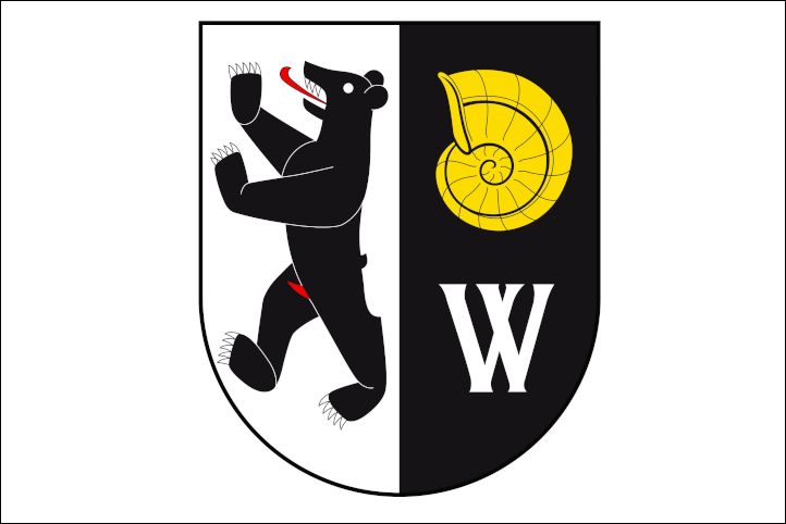 Das Bild zeigt ein Wappen, das auf der linken Seite einen aufrechten, schwarzen Bären mit rotem Maul und auf der rechten Seite eine gelbe Schnecke zeigt. Unten ist ein großes, weißes "W" zu sehen. Der Hintergrund ist in schwarz und weiß geteilt.