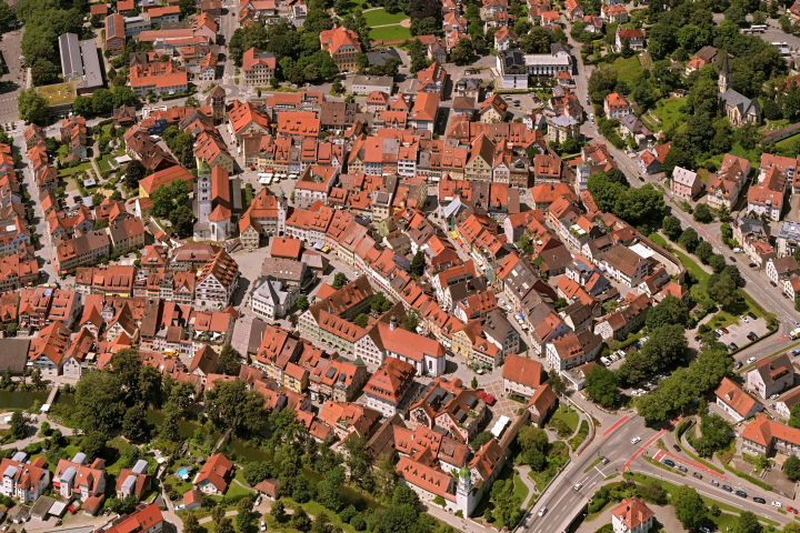 Luftaufnahme einer Stadt mit vielen Häusern und roten Dachziegeln, umgeben von grünem Gelände. Die Straßen sind gut zu erkennen, und es gibt verschiedene Gebäudetypen, die eine historische Architektur vermuten lassen. Ein Platz ist in der Mitte sichtbar, umgeben von Bäumen und Parklandschaft.