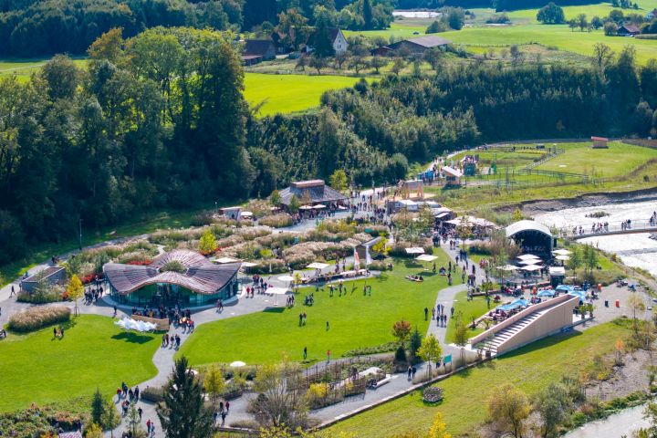 Eine Luftaufnahme eines Parks mit vielen Besuchern, die sich auf Grünflächen und Wegen verteilen. Es sind verschiedene Aktivitäten und Stände sichtbar, umgeben von Bäumen und Hügeln. Im Hintergrund sind landwirtschaftliche Flächen und Gebäude zu erkennen.