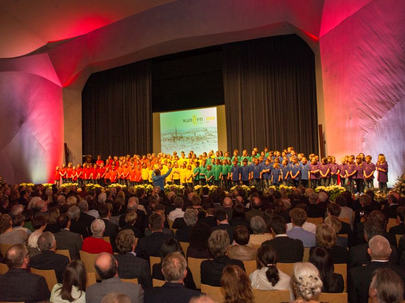 Ein großer Kinderchor, bekleidet in T-Shirts in Regenbogenfarben, singt im voll besetzten Festsaal der Waldorfschule
