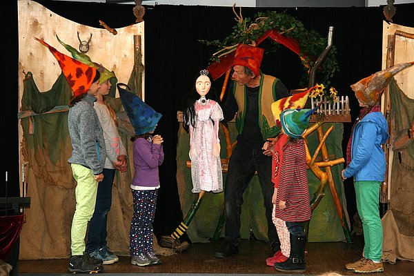 Eine Gruppe von Kindern in bunten Hüten steht auf einer Bühne. Ein Erwachsener, ebenfalls mit einem bunten Hut, präsentiert eine Puppe. Im Hintergrund sind zwei große, dekorierte Banner zu sehen, die zur Theateraufführung gehören. Die Atmosphäre ist lebhaft und fröhlich.