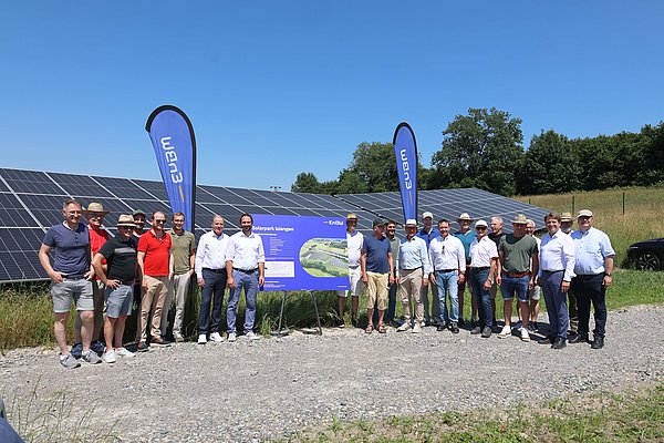 Gruppe von Menschen, die sich vor einer Solaranlage versammelt haben. Im Hintergrund sind große Solarpanels und ein Banner sichtbar. Der Himmel ist blau und es herrscht sonniges Wetter. Die Personen tragen unterschiedliche Kleidung und einige haben Hüte auf.