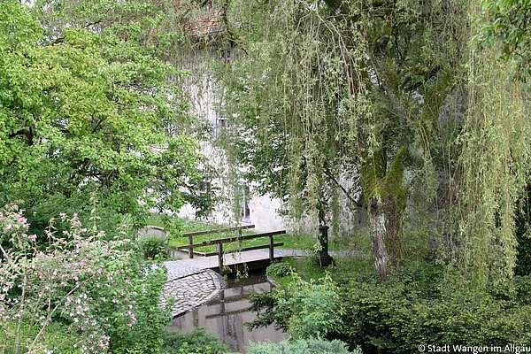 Eine kleine Holzbrücke über einen schmalen Wasserlauf ist von dichtem Grün umgeben. Hohe Bäume, Sträucher und Pflanzen sorgen für eine natürliche Atmosphäre, während im Hintergrund ein Gebäude zu sehen ist.