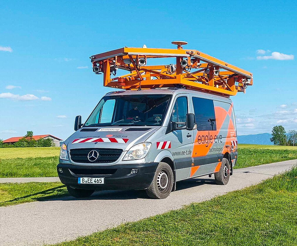 Ein grauer Lieferwagen mit orangefarbener Ausrüstung auf dem Dach steht auf einer ländlichen Straße. Umgeben von grünen Wiesen und einem blauen Himmel mit leichten Wolken.