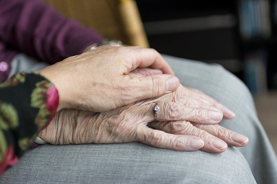 Zwei Hände liegen übereinander, eine älter und mit sichtbaren Falten, die andere jünger und zärtlich darauf platziert. Die ältere Hand trägt einen schlichten Ring. Der Hintergrund ist unscharf, mit einem warmen, häuslichen Ambiente.