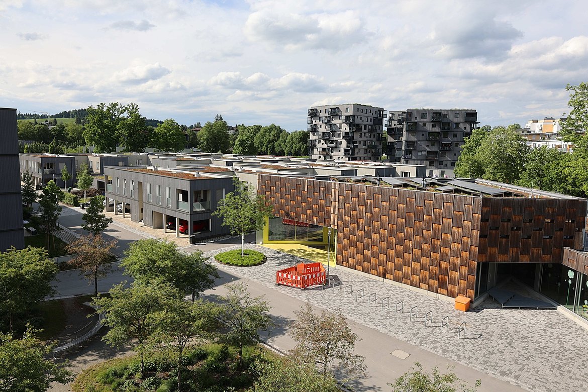 Moderne Architektur in einer städtischen Umgebung. Es sind mehrere Gebäude sichtbar, einige mit Holzverkleidung, andere in grauem Beton. Im Vordergrund gibt es grüne Landschaftselemente und einen gepflasterten Bereich. Der Himmel ist teilweise bewölkt.