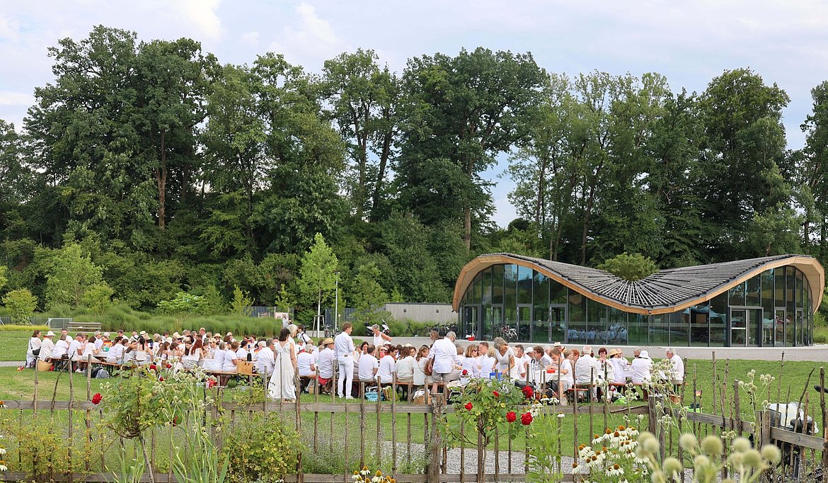Eine große Gruppe von Menschen in weißer Kleidung sitzt auf Bänken und an weiß-gedeckten Tischen auf einer Wiese vor einem modernen Gebäude mit einer gewellten Dachkonstruktion. Im Hintergrund sind Bäume und ein gepflegter Garten sichtbar.