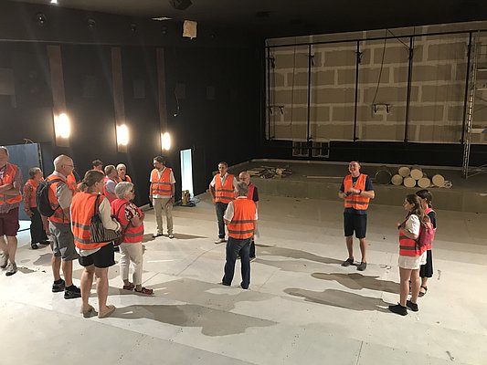 Eine Gruppe von Personen in orangefarbenen Warnwesten steht in einem großen, leeren Raum. Einige Menschen sprechen miteinander, während andere aufmerksam zuhören. Die Umgebung hat eine dunkle Wandfarbe und es gibt einige Lichter an der Decke.