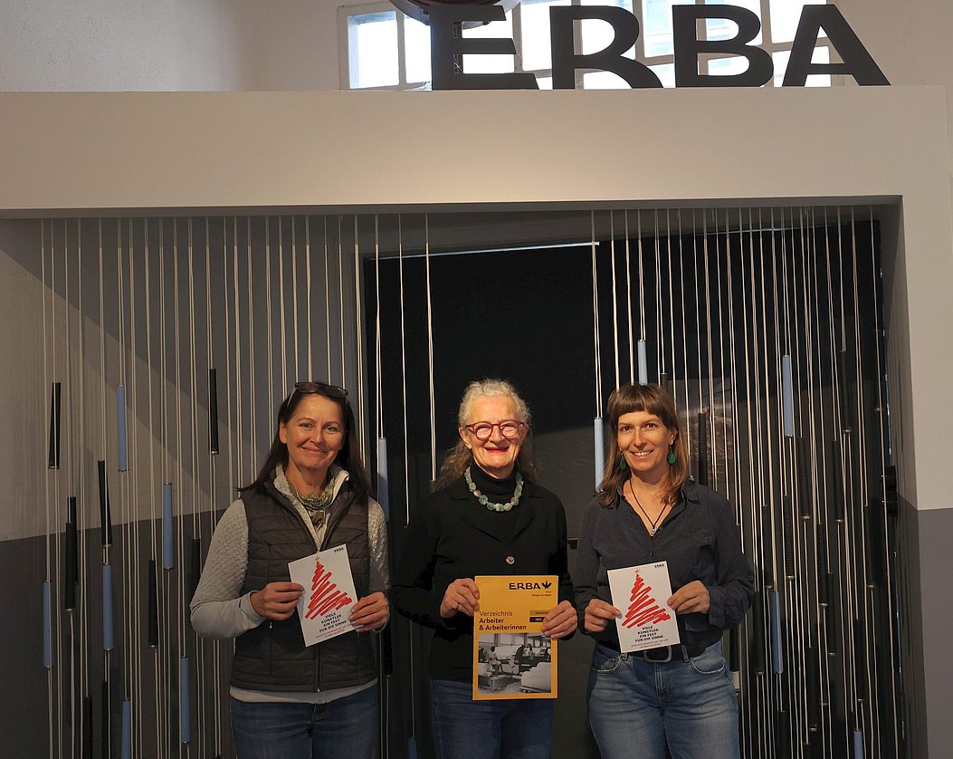 3 Frauen. Die Frau in der Mitte hält einen Bucheinband, die beiden links und rechts einen Zettel mit einem stilisierten Weihnachtsbaum.