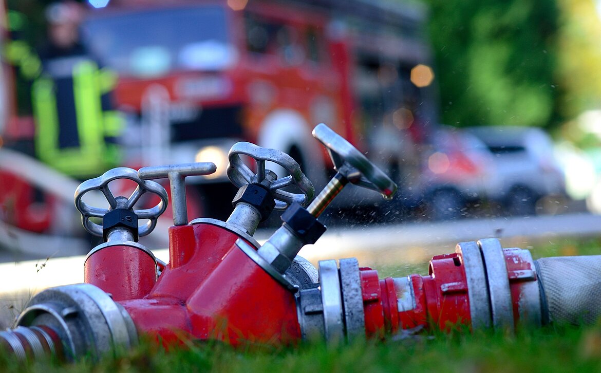 Ein roter Feuerwehrhydrant liegt auf dem Boden, mit zwei Ventilen sichtbar. Im Hintergrund ist ein Feuerwehrfahrzeug und ein Feuerwehrmann zu erkennen. Wassertropfen spritzen von der Düse des Hydranten.