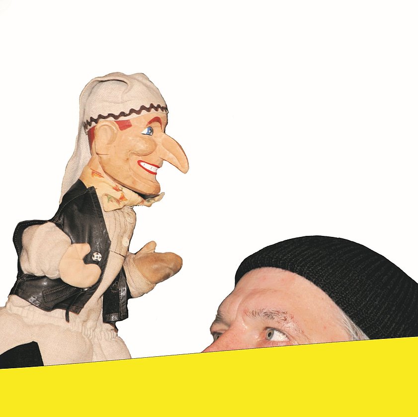 Ein Handpuppenspieler mit einer Puppe, die eine Kasperlfigur darstellt. Die Puppe hat eine große, orangefarbene Nase und trägt ein Kasperlkostüm. Der Puppenspieler trägt eine schwarze Kappe  und schaut aufmerksam auf die Puppe. Der Hintergrund ist hell und einfarbig.