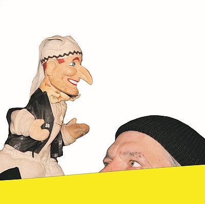 Ein Handpuppenspieler mit einer Puppe, die eine Kasperlfigur darstellt. Die Puppe hat eine große, orangefarbene Nase und trägt ein Kasperlkostüm. Der Puppenspieler trägt eine schwarze Kappe  und schaut aufmerksam auf die Puppe. Der Hintergrund ist hell und einfarbig.