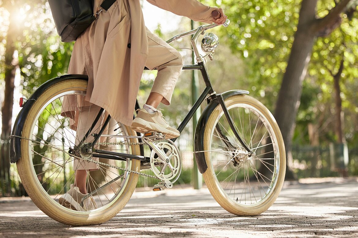 Eine Person sitzt auf einem Fahrrad mit einem schwarzen Rahmen und breiten, hellem Reifen. Sie trägt bequeme Kleidung und Sneakers. Im Hintergrund sind grüne Bäume und Lichtschimmer zu sehen, die eine entspannte, urbane Umgebung schaffen.