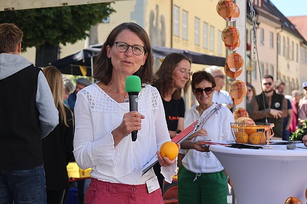 Eine Frau mit Brille hält ein Mikrofon mit grünem Kopf und eine Orange in der Hand. Sie trägt eine weiße Bluse und pinkfarbene Hose. Im Hintergrund sind weitere Personen und Tische mit Obst und Notizen zu sehen, während die Veranstaltung im Freien stattfindet.