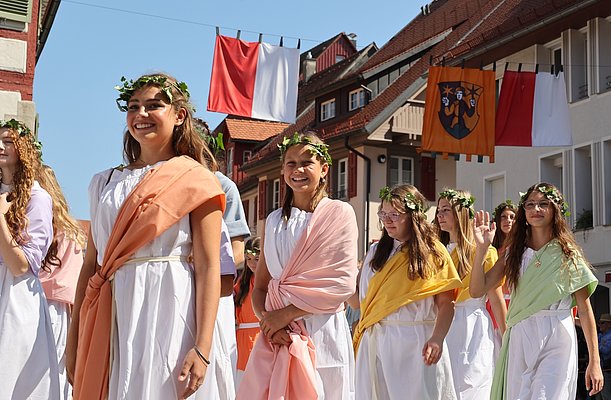 Junge Frauen in weißen Gewändern mit bunten Schärpen und Blumenkränzen auf dem Kopf lächeln während einer Festparade. Sie gehen auf einer Straße, umgeben von traditionellen Gebäuden und Fahnen. Die Szenerie strahlt Festlichkeit und Freude aus.