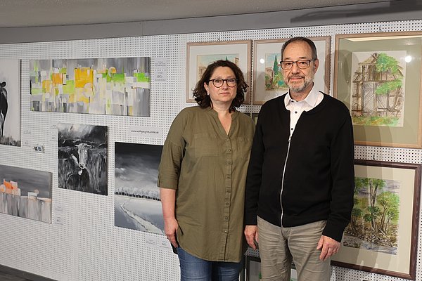 Ein Mann und eine Frau stehen vor einer Wand mit verschiedenen Kunstwerken. Die Wand zeigt Gemälde und Zeichnungen in unterschiedlichen Stilen und Farben, darunter Landschaften und abstrakte Arbeiten. Die beiden Personen lächeln und wirken freundlich.