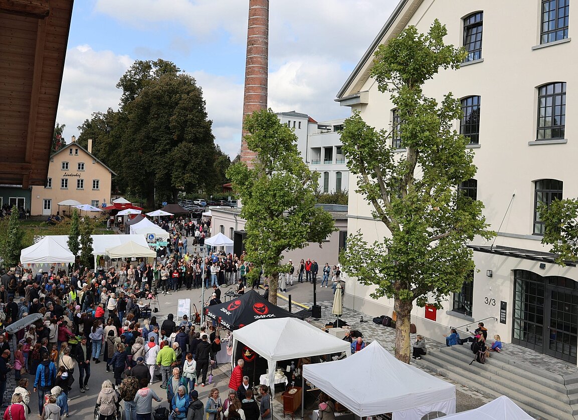 Eine große Menschenmenge versammelt sich auf einem Platz, umgeben von Ständen mit Zelten und Bäumen. Im Hintergrund sind zwei Gebäude sichtbar, darunter ein hoher Schornstein. Der Himmel ist teilweise bewölkt.