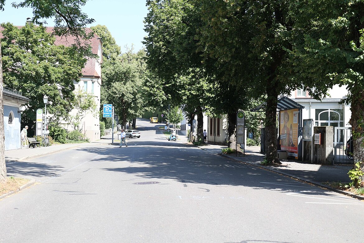 Eine ruhige Straße mit Bäumen an beiden Seiten. Auf der linken Seite sind eine Parkhausbeschilderung und eine Wartebank. Fahren mehrere Fahrzeuge, darunter ein gelbes Auto. Es sind einige Fußgänger unterwegs.