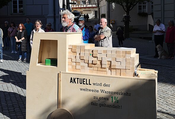 Eine Gruppe von Menschen steht auf einem Platz. Im Vordergrund ist eine große Holzstruktur sichtbar, die mit Blöcken gefüllt ist und einen Aufruf zur fairen Handelsvertretung enthält. Im Hintergrund sind weitere Passanten zu sehen.