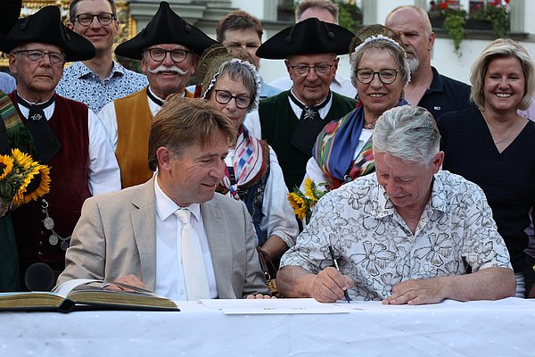 Zwei Männer sitzen an einem Tisch und unterzeichnen Dokumente. Im Hintergrund stehen mehrere Personen in traditioneller Kleidung, die Sonnenblumen halten. Die Szene wirkt feierlich und festlich.