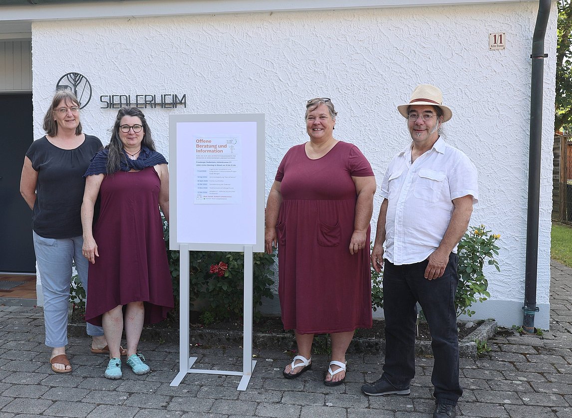 Vier Personen stehen vor einem Gebäude mit dem Schriftzug "Siedlerheim". Sie tragen ähnliche rot- und blaufarbene Kleider und lächeln. Neben ihnen steht ein Informationsschild, das offenbar Veranstaltungshinweise enthält. Der Hintergrund zeigt eine weiße Wand und einen gepflasterten Boden.