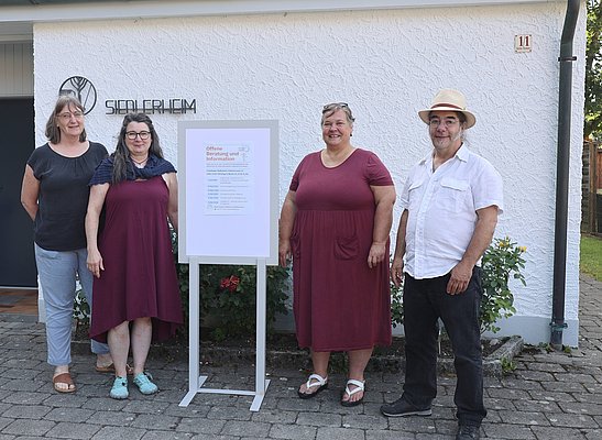 Vier Personen stehen vor einem Gebäude mit dem Schriftzug "Siedlerheim". Sie tragen ähnliche rot- und blaufarbene Kleider und lächeln. Neben ihnen steht ein Informationsschild, das offenbar Veranstaltungshinweise enthält. Der Hintergrund zeigt eine weiße Wand und einen gepflasterten Boden.