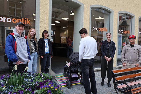 Mehrere Personen beobachten einen jungen Mann. Er schiebt einen Kinderwagen über eine Rampe.